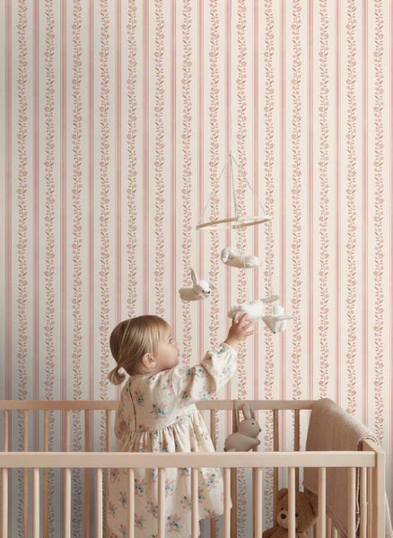 Ein Kleinkind in einem hölzernen Babybett greift nach einem Mobile vor einer Kinderzimmertapete mit Streifen und Blumenranken.
