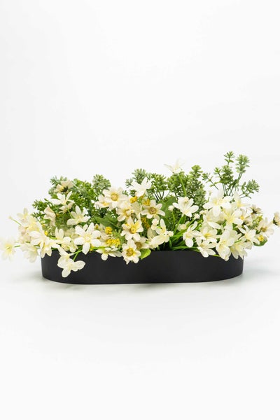 Künstliches Blumengesteck mit weißen Blüten und grünen Blättern in einer ovalen schwarzen Schale.