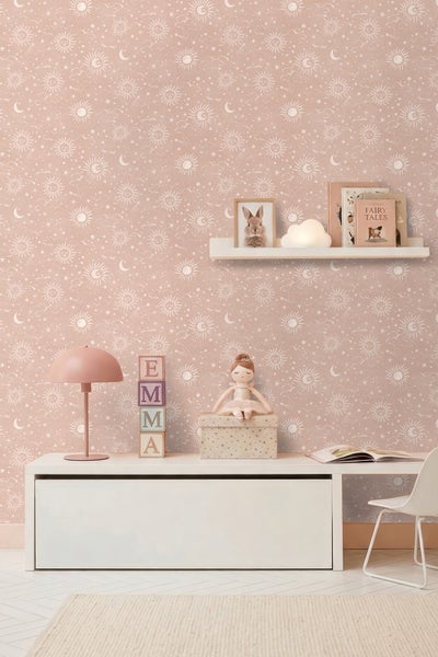 Rosa Kinderzimmer-Tapete mit Sonnen-, Mond- und Sternenmuster in einem hell eingerichteten Raum mit weißen Möbeln.