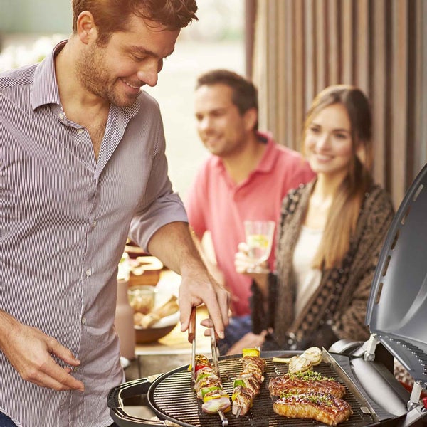 Szene mit Grill und grillendem Mann, während zwei Personen im Hintergrund sitzen