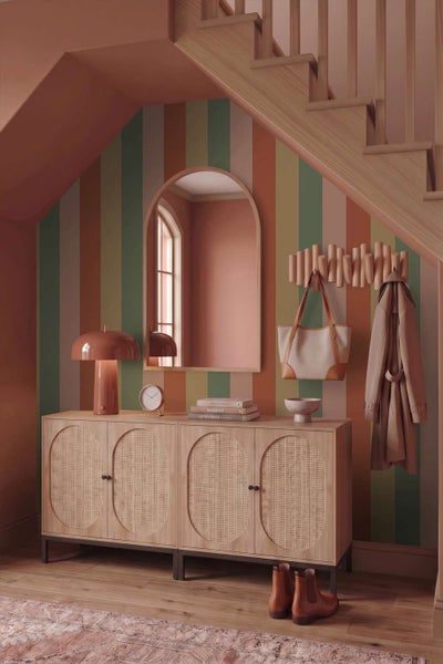 Flur mit gestreifter Tapete, Sideboard mit Geflechtfronten, Bogenspiegel, Kleiderhaken und Tischlampe unter einer Holztreppe.