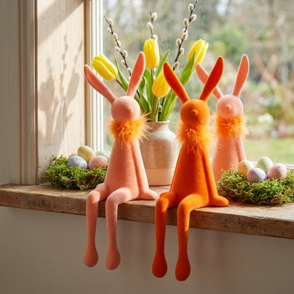 Drei sitzende Osterhasen aus Stoff mit Federkragen als Tischdekoration auf einer Fensterbank mit Frühlingsblumen und Ostereiern.