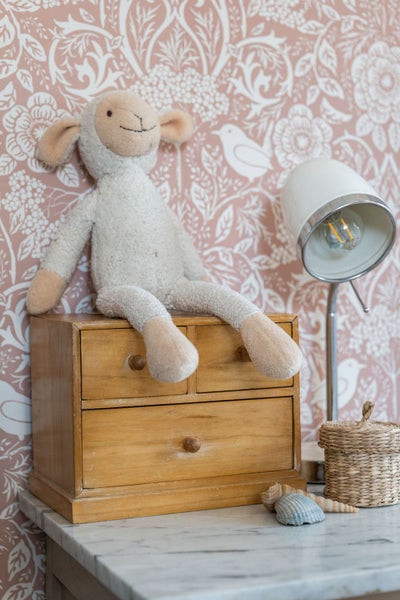 Kinderzimmerdekoration mit gemusterter Tapete, kleiner Kommode aus Holz, Plüschschaf, Tischleuchte, Flechtkorb und Muscheln auf einer Marmoroberfläche.