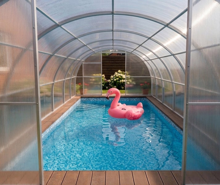 Überdachter Gartenpool mit aufblasbarem Flamingo und Holzterrasse in einer hellen, wettergeschützten Schwimmbadüberdachung aus Polycarbonat.