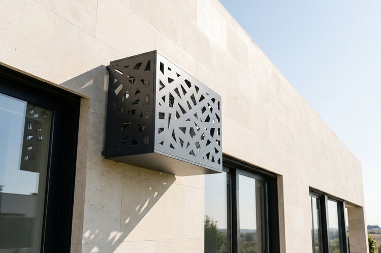 Modern gestaltete Klimaanlagen-Abdeckung aus Metall mit geometrischem Muster an einer hellen Steinfassade neben Fenstern.