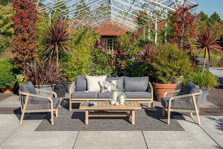 Modernes Garten-Lounge-Set aus Holz mit Sofa, Sesseln und Tisch auf einer Terrasse, umgeben von üppigen Pflanzen in einem hellen Garten.