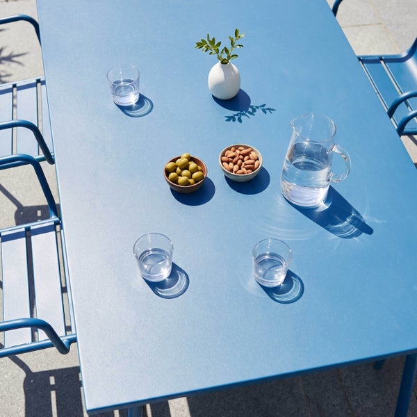 Rechteckiger blauer Metall-Gartentisch mit passenden Metallstühlen, gedeckt mit einer Glaskaraffe, Wassergläsern, Oliven und Mandeln im Freien.