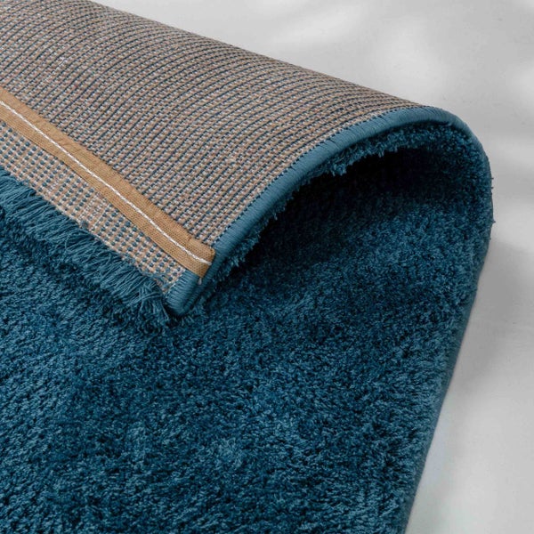 Nahaufnahme eines petrolfarbenen Hochflor-Teppichs mit weicher Textur und gewebter Rückseite.