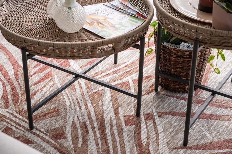 Outdoor-Teppich mit abstraktem Muster unter runden Beistelltischen aus Rattangeflecht mit Metallgestell.