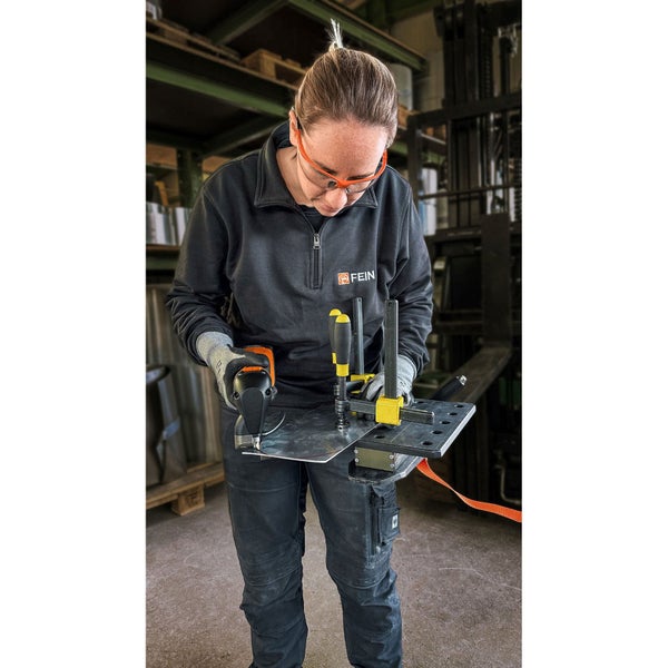 Frau schneidet Metallplatte mit einem Fein Knabber in einer Werkstatt, trägt Schutzkleidung und Schutzbrille.
