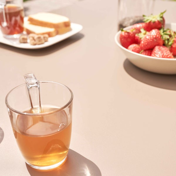 Nahaufnahme eines Teeglases auf einem Gartentisch mit Erdbeeren und Brot im Hintergrund an einem sonnigen Tag.
