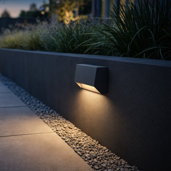 Moderne LED-Außenwandleuchte an einer Gartenmauer, die einen Weg aus Kies und Steinplatten mit warmweißem Licht beleuchtet.