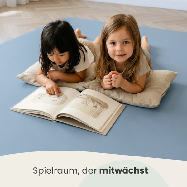 Zwei Kinder liegen auf einer blauen Spielmatte und beigen Kissen und lesen ein Buch in einem hellen Raum mit hellem Holzboden.