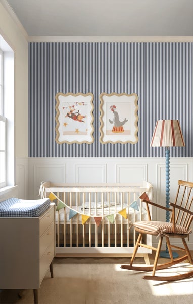 Helles Kinderzimmer mit blau-weiß gestreifter Tapete, weißer Wandtäfelung, Gitterbett aus Holz, Schaukelstuhl und Bildern mit Zirkusmotiven.
