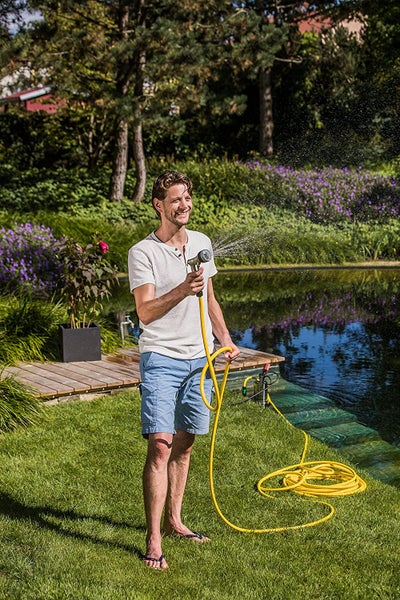 Ein Mann bewässert einen sonnigen Garten mit einem gelben Gartenschlauch und einer Spritzdüse neben einem Gartenteich mit Holzsteg.