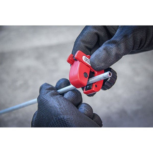 Roter Mini-Rohrabschneider beim Schneiden eines Metallrohrs, gehalten von Händen in schwarzen Handschuhen.