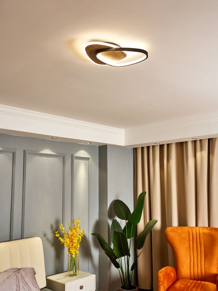 Moderne LED-Deckenleuchte mit zwei überlappenden Ringen in Holzoptik in einem hellen Wohnraum mit grauen Wänden und beigen Vorhängen.