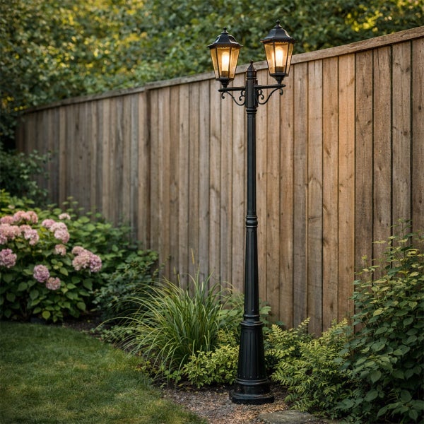 Zweiflammige Gartenlaterne aus schwarzem Metall im klassischen Design vor einem Holzzaun in einem bepflanzten Garten.