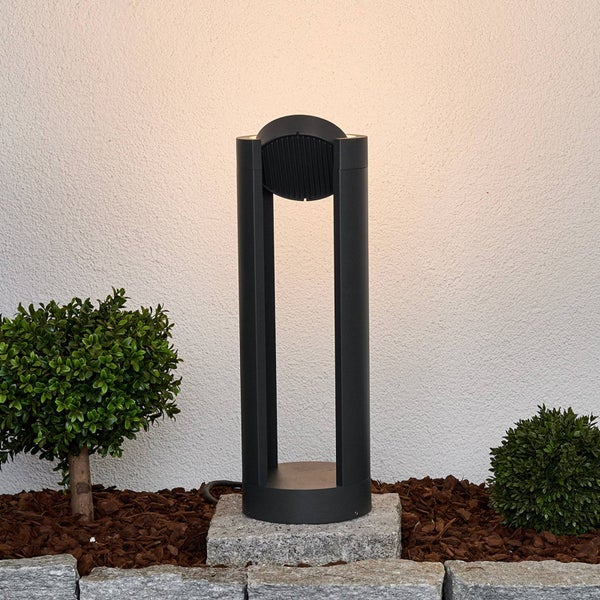 Moderne Pollerleuchte für den Garten, anthrazitfarbenes Design auf Steinsockel in Beet mit Rindenmulch vor einer hellen Wand.