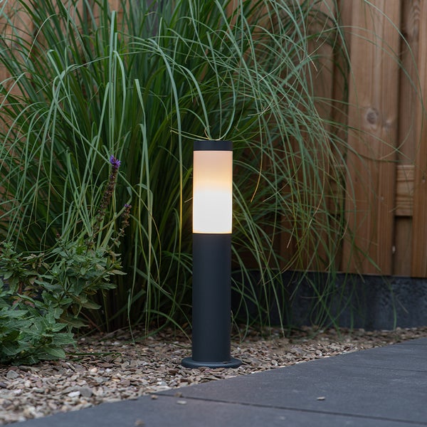 Moderne LED Außen-Wegeleuchte in Anthrazit, platziert in einem Gartenbeet vor Gräsern und einem Holzzaun.
