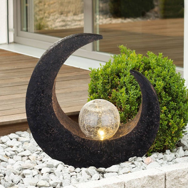 Solar-Gartenleuchte in Mondsichel-Optik mit Glaskugel in Bruchglas-Design auf hellem Kies neben einem grünen Strauch.