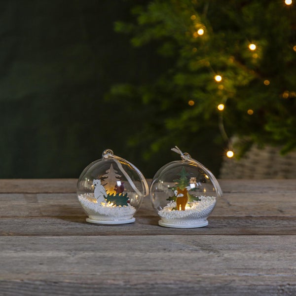 Zwei durchsichtige Weihnachtskugeln mit Waldtiermotiven und Kunstschnee auf einem Holztisch