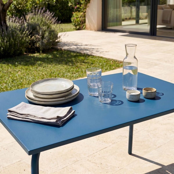 Gedeckter blauer Metall-Gartentisch auf einer Terrasse mit Keramiktellern, Stoffservietten und Gläsern vor einem sonnigen Garten.