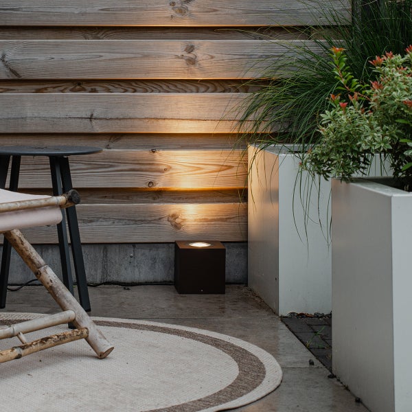 Quadratische Außenbodenleuchte beleuchtet eine Holzwand auf einer Terrasse mit Pflanzgefäßen, einem Teppich und einem Tisch.
