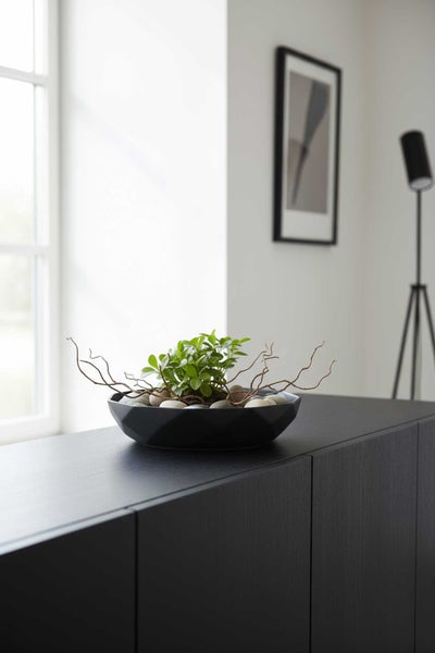 Schwarze geometrische Dekoschale mit grüner Pflanze, Steinen und Zweigen auf einem dunklen Sideboard.