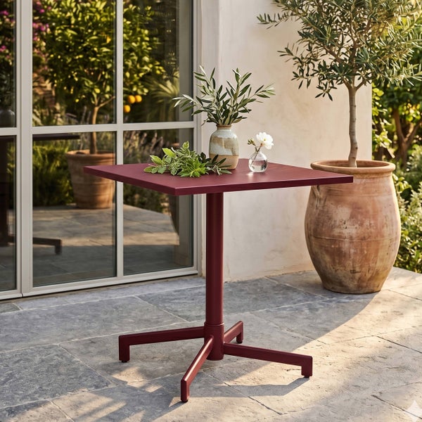 Quadratischer Bistro-Tisch aus rotem Metall auf einer Steinterrasse mit dekorativen Vasen und einem Olivenbaum im Hintergrund.