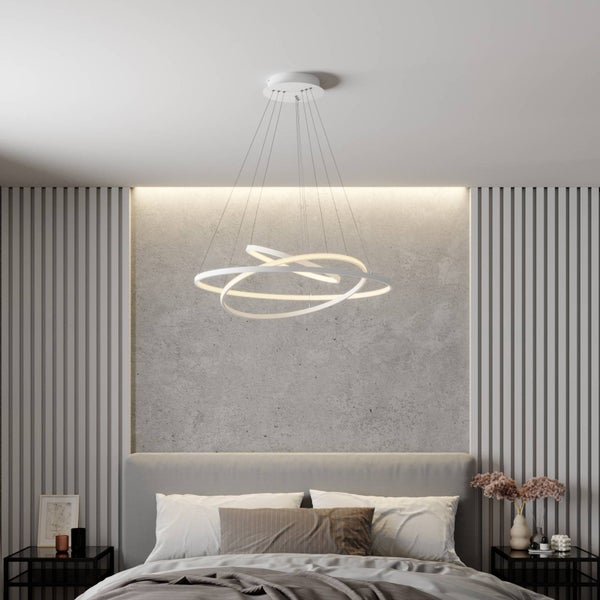 Moderne Schlafzimmer-Szene mit einer weißen LED-Pendelleuchte aus drei Ringen über einem grauen Bett vor einer Wand in Betonoptik.