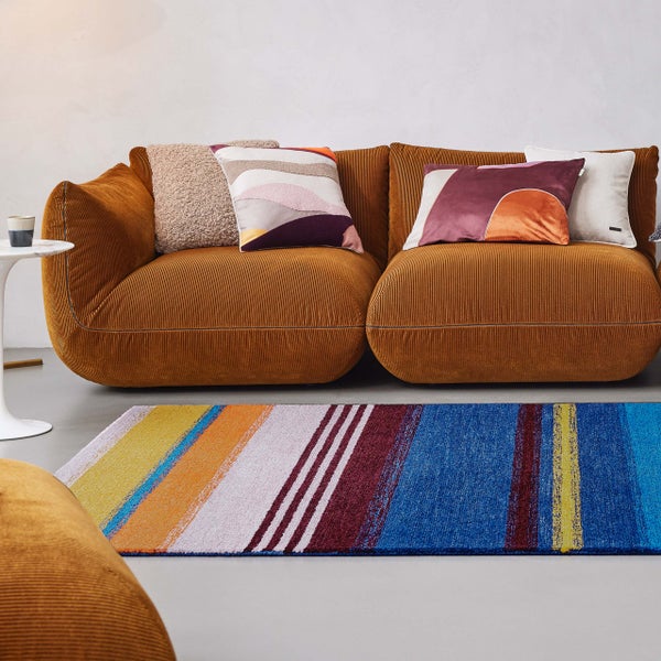 Modernes Wohnzimmer mit braunem Cord-Sofa, bunten Dekokissen und einem mehrfarbig gestreiften Teppich.