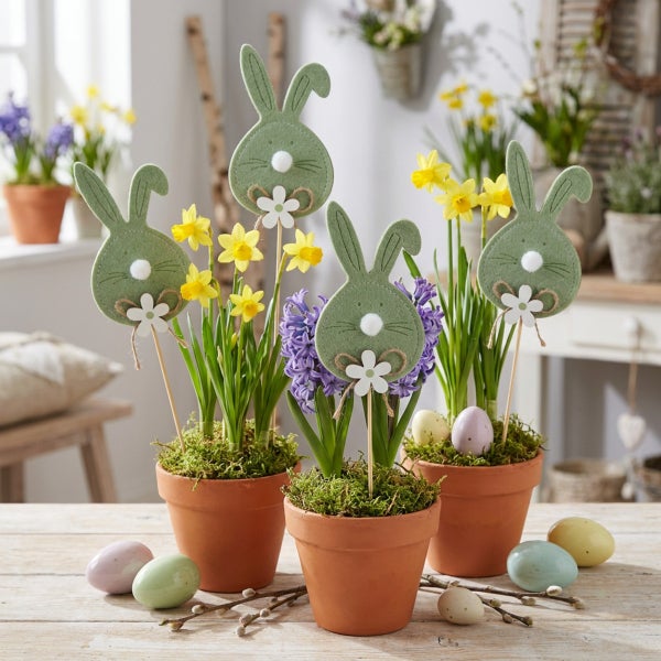 Drei Terrakottatöpfe mit Frühlingsblumen wie Narzissen und Hyazinthen, dekoriert mit Filzhäschen auf Holzstäben und verstreuten Ostereiern.