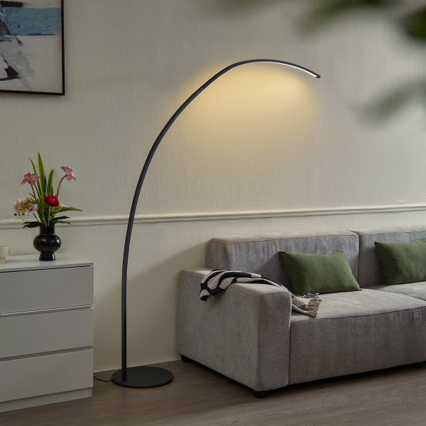 Moderne schwarze LED-Bogenstehlampe mit gebogener Lichtquelle neben einem grauen Sofa in einem hellen Wohnzimmer.