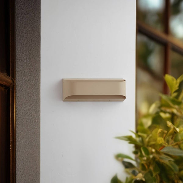 Moderne beige rechteckige Außenwandleuchte mit abgerundeten Kanten, montiert an einer Außenwand neben einem Fenster und Pflanzen.
