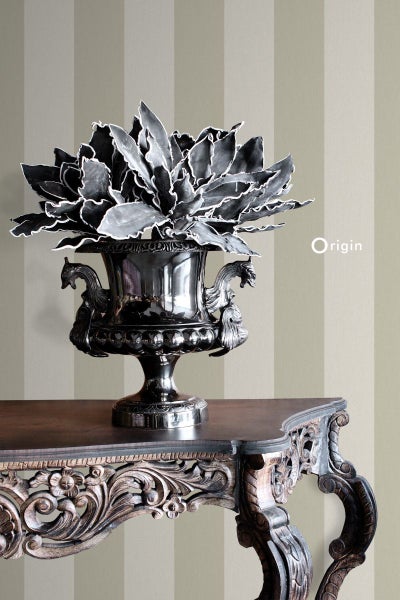 Dekorative Kunstblume in silberner Vase auf einem verzierten Holztisch vor einer gestreiften Tapete.