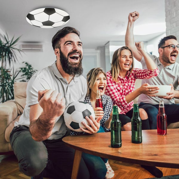 Eine runde LED Deckenleuchte im Fußball Design leuchtet in einem Wohnzimmer, in dem eine Gruppe Menschen begeistert ein Fußballspiel verfolgt.