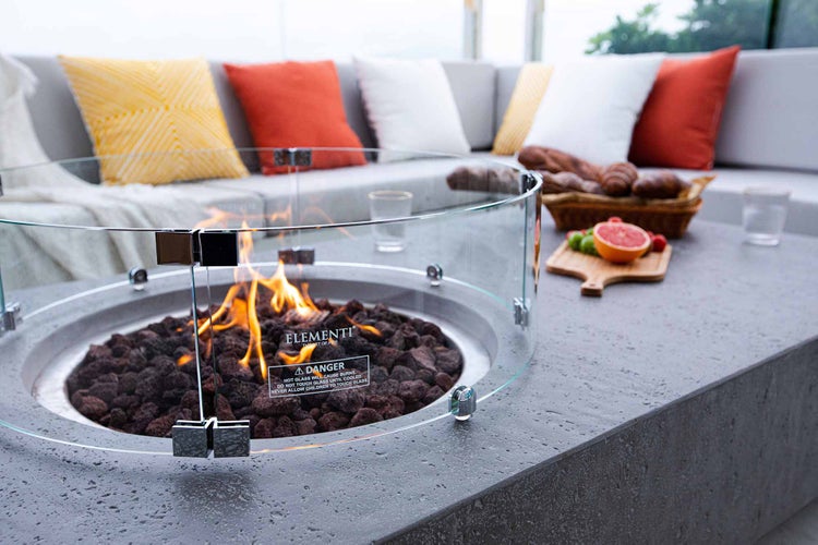 Moderner Gas-Feuertisch mit rundem Glasschutz und Lavasteinen auf einer Terrasse mit Loungemöbeln.