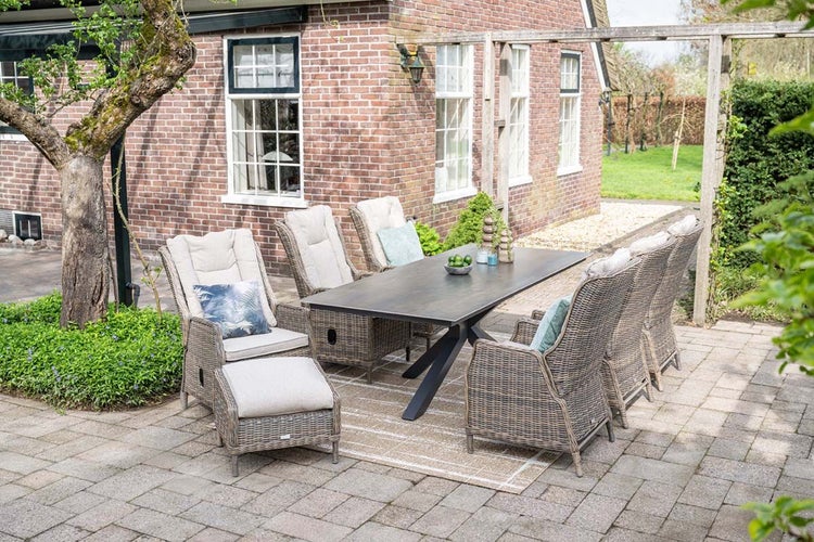 Garten-Essgruppe auf gepflasterter Terrasse, bestehend aus einem dunklen Esstisch und sechs Polyrattan-Sesseln mit hellen Kissen und Fußhocker.