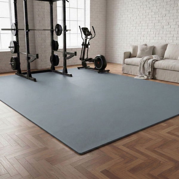Große graue Bodenschutzmatte für Fitnessgeräte in hellem Raum mit Hantelstation, Crosstrainer und Sofa auf Parkettboden.