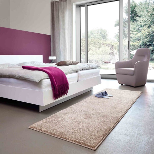 Modernes helles Schlafzimmer mit weißem Bett, lila Wand, grauem Boden und beigem Teppichläufer vor einem großen Fenster mit Blick in den Garten.