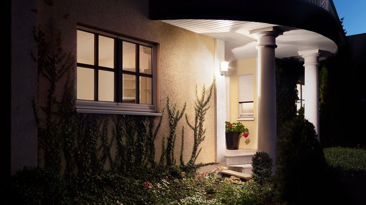Beleuchtete Hausfassade bei Nacht mit Außenwandleuchte, Fenster, Kletterpflanzen und Säulen im Eingangsbereich.