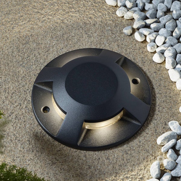 Runder, schwarzer Bodenstrahler für außen, eingebaut in Steinboden, seitlicher Lichtaustritt neben hellen Kieseln.