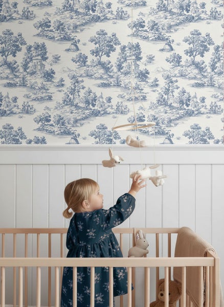 Helles Kinderzimmer mit Toile-de-Jouy-Tapete und weißer Wandvertäfelung aus Holz neben einem hölzernen Gitterbett.