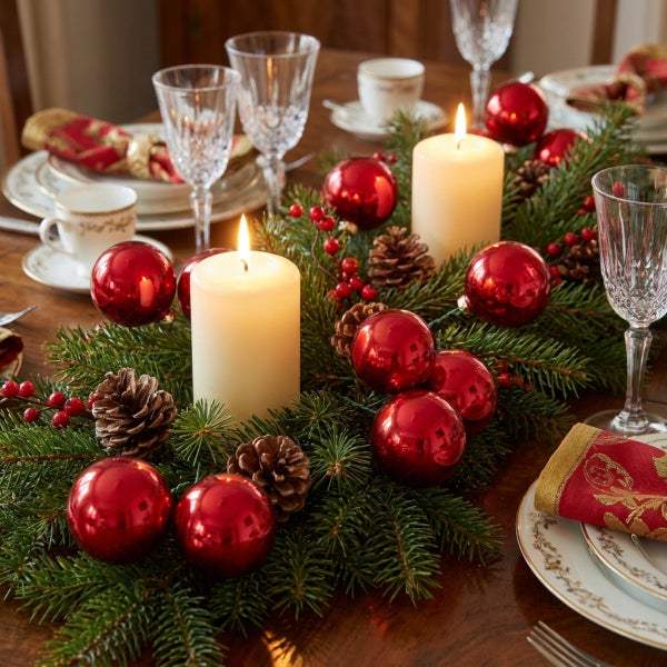 Festliche Tischdekoration mit Tannenzweigen, brennenden Stumpenkerzen, roten Weihnachtskugeln, Zapfen und Beerenzweigen auf einem Holztisch.