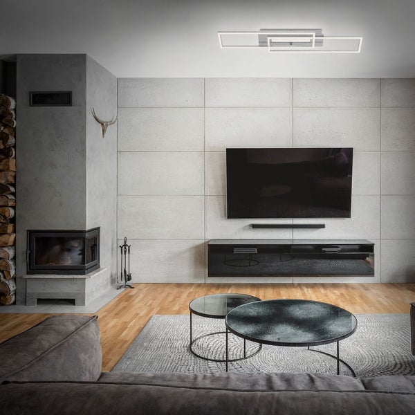 Modernes Wohnzimmer mit geometrischer LED-Deckenleuchte, TV an Betonoptik-Wand, grauem Kamin und runden Couchtischen auf Holzboden.