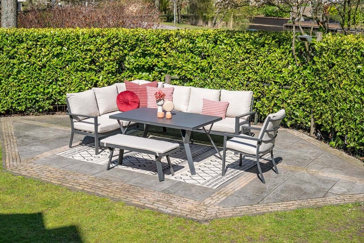 Modernes Gartenmöbel Set mit Ecksofa, Esstisch, Bank und Sessel auf einer Steinterrasse vor einer Hecke.