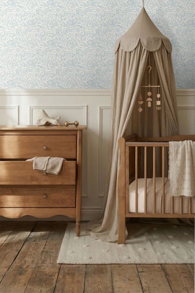 Helles Babyzimmer mit blau-weißer Blumentapete, Wandpaneelen, einer Holzkommode und einem Kinderbett aus Holz mit beigem Baldachin.