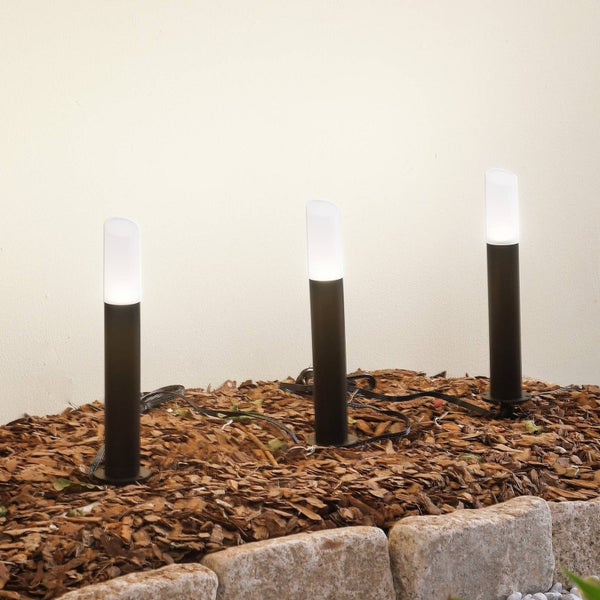 Drei schwarze zylindrische Gartenleuchten in einem mit Rindenmulch bedeckten Beet mit Steineinfassung und weißem Kies. Helle Außengestaltung.