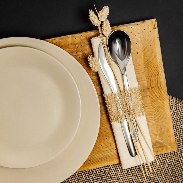 Gedeckter Tisch mit Keramiktellern, Holzbrett, Edelstahlbesteck und Leinenserviette mit Juteband und Trockenblumen.
